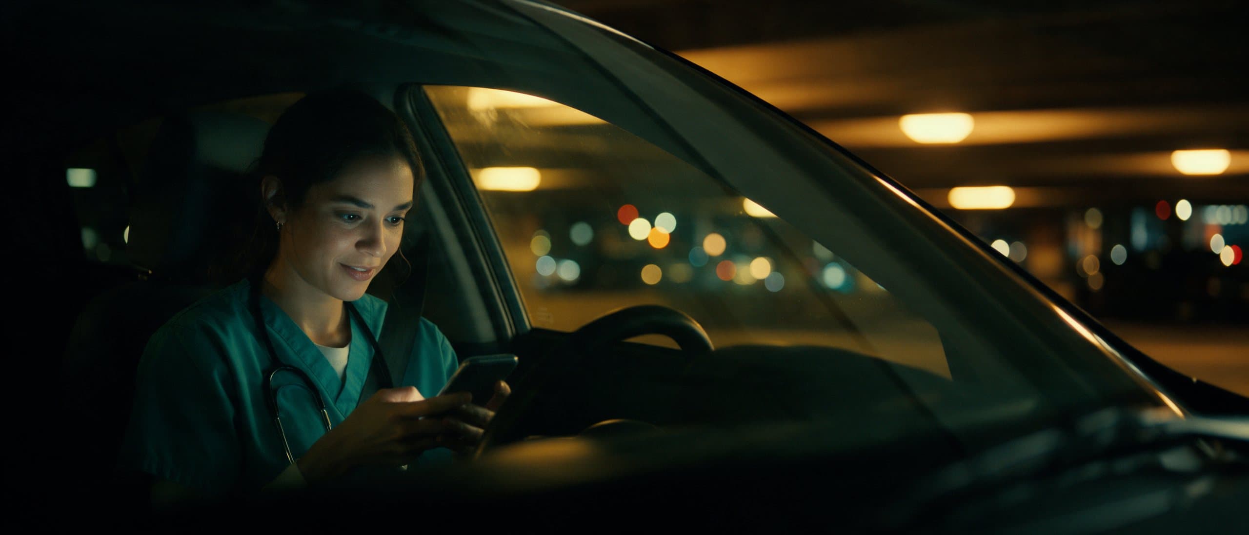 A technologist in scrubs typing on her phone in a parked car after a shift