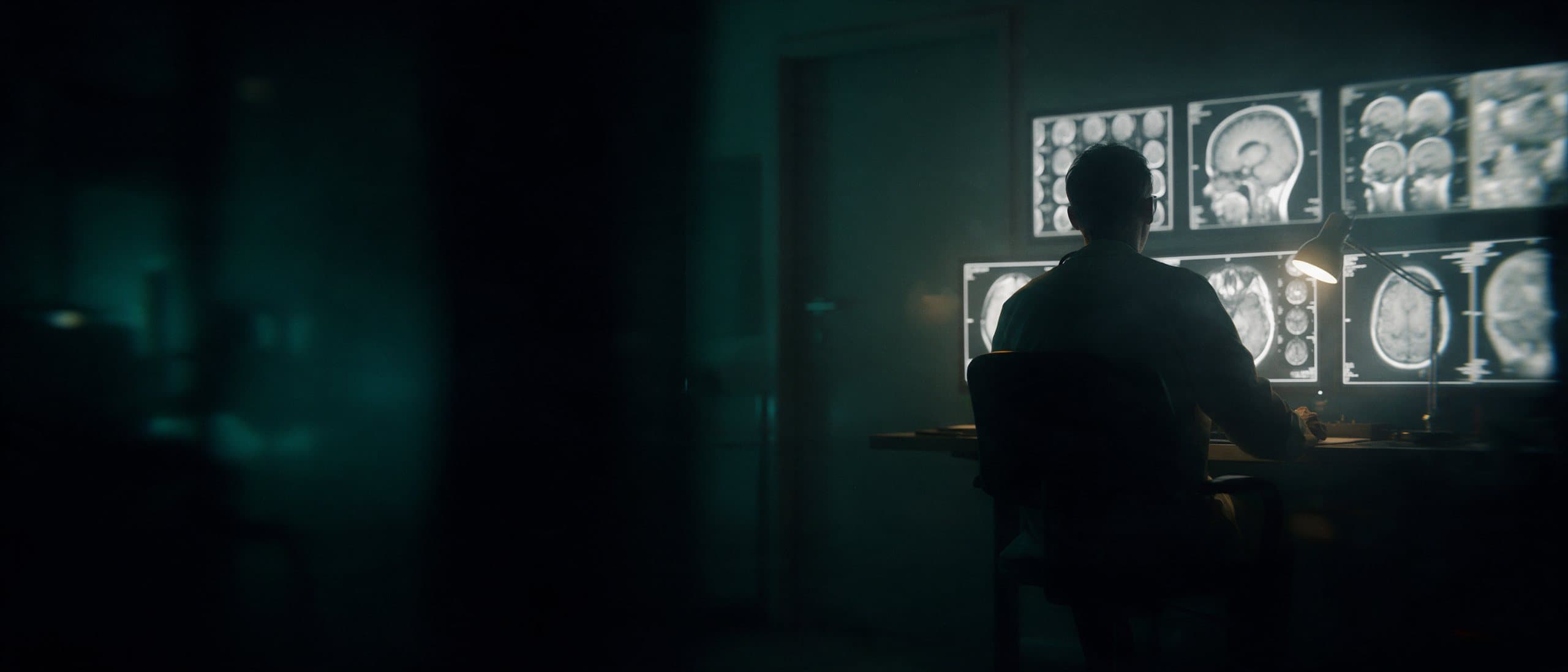 A radiologist alone in a dark reading room lit by scan monitors